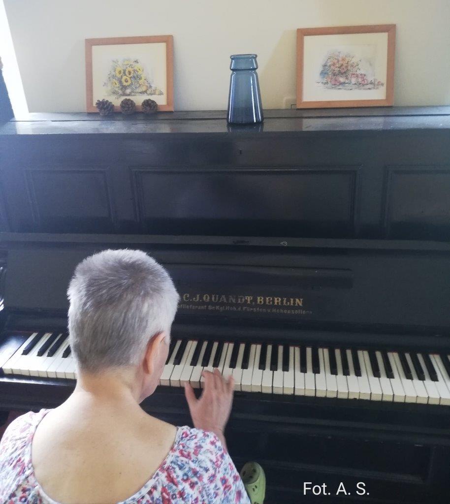  Zdjęcie kobiety grającej na czarnym pianinie. na pianinie stoi niebieski wazon obok dwa obrazki z namalowanymi kwiatami , w tle beżowa ściana