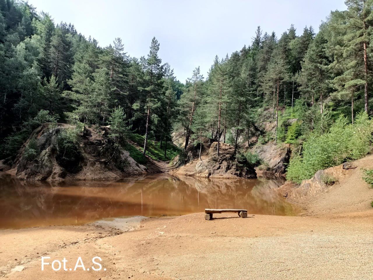 Zdjęcie jeziorka w kolorze  purpurowym, dookoła widać strome zbocza porośnięte drzewami iglastymi, na dole zdjęcia kamienista plaża z ławeczką z bali. Ponad linią drzew jasno błękitne niebo