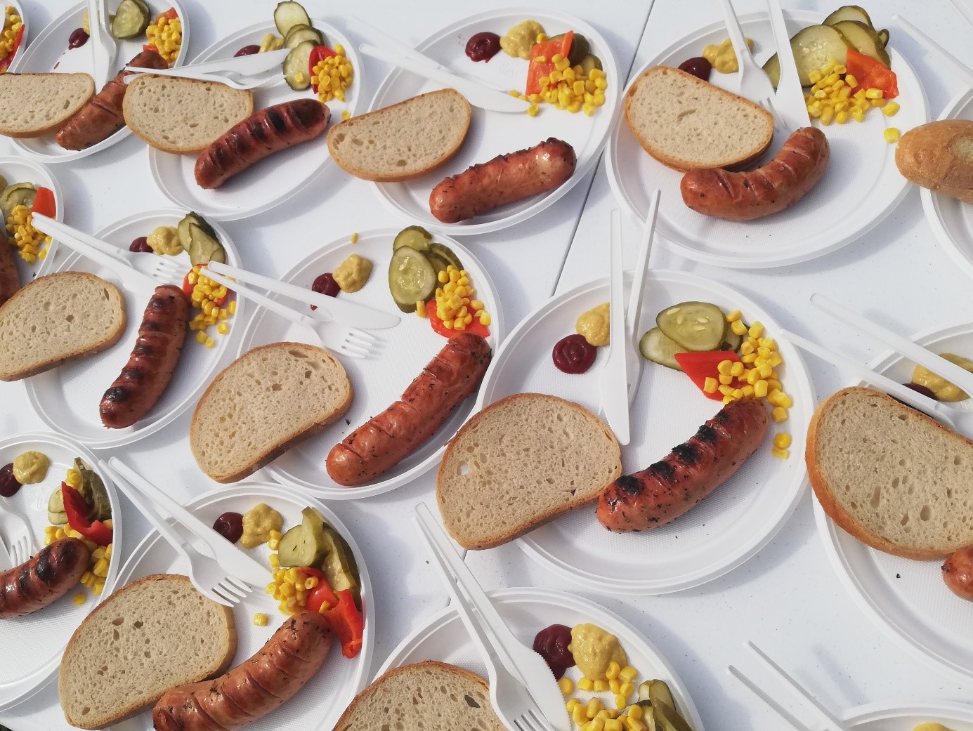 Na zdjęciu na stole z białym obrusem, kilkanaście białych talerzy ułożonych obok siebie. Na Każdym talerzu grillowa kiełbasa, kromka chleba, musztarda, ketchup i mieszanka warzywna: kukurydza, papryka, ogórek oraz plastikowy widelec i nóż 