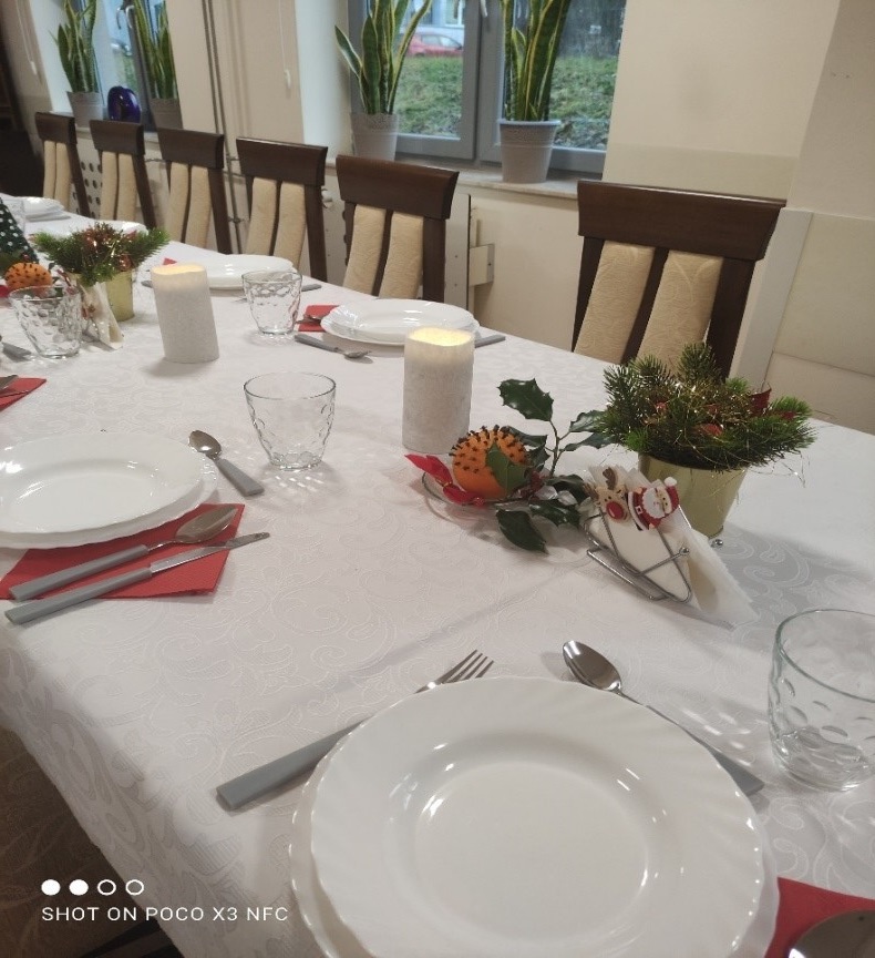  Aranżacja stołu wigilijnego z białym obrusem i dodatkami ze świątecznymi motywami. Zastawa stołowa w kolorze białym, komplet sztućców, świece, pomarańcze z goździkami, stroiki świąteczne.
