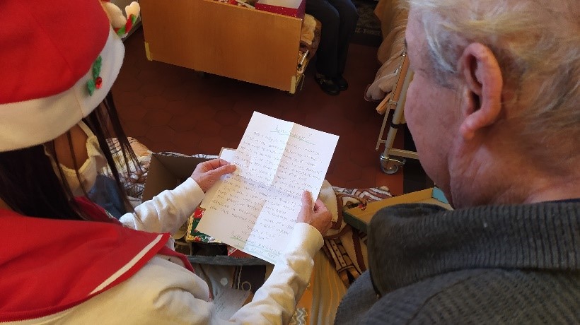 Na zdjęciu kobieta i mężczyzna wspólnie czytają list. W tle brązowa podłoga i fragment stojącego na niej łóżka