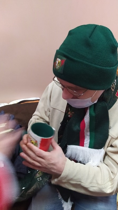 Na zdjęciu mężczyzna ubrany w jasną koszulę z długim rękawem, na głowie zielona czapka, na szyi zielono-biało-czerwony szalik. W ręku kubek w kolorach zielono-biało-czerwony (gadżety Wrocławskiego Klubu Sportowego