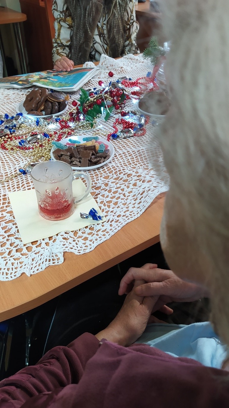 Na zdjęciu kobiety siedzące przy brązowym stole z białym szydełkowym obrusem. Na stole ciastka, cukierki, kubek z napojem oraz świąteczne ozdoby (koralikowy łańcuch, zielona gałązka, światełka)