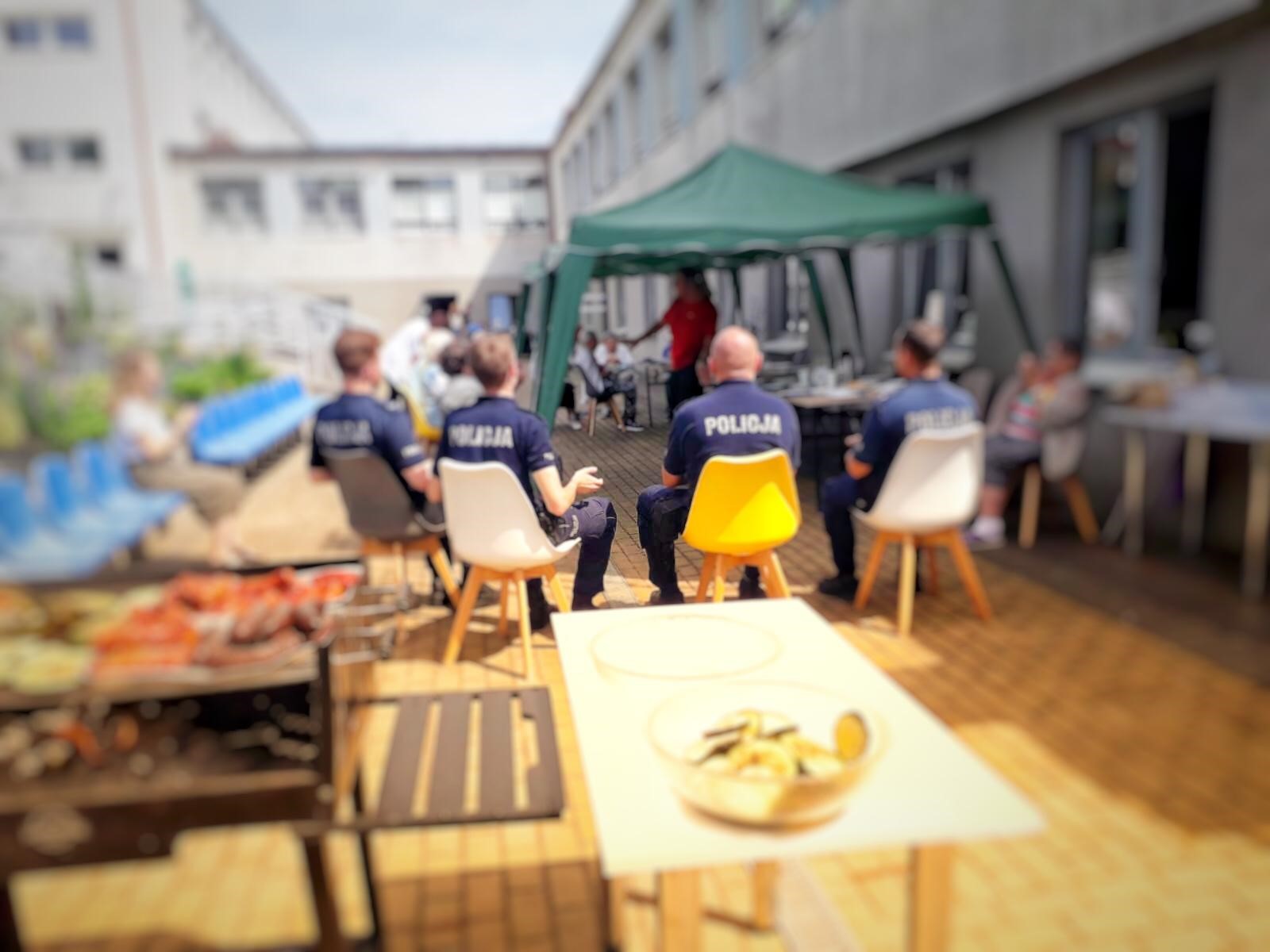 Teren zewnętrzny DDP. Na grillu smaży się mięso oraz warzywa, obok grilla stoi biały stół. Na kolorowych krzesłach siedzi 4 policjantów. Na dalszym planie widoczne są zielone namioty ochraniające przed słońcem, na krzesłach zaś siedzą Klienci DDP. Wszyscy biorą udział w grupowej gimnastyce