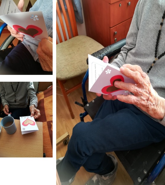 Mieszkanka DPS siedzi na wózku inwalidzkim, ogląda kartkę otrzymaną z okazji obchodów Międzynarodowego Dnia Chorego.  Kartka, wykonane z białego, gładkiego papieru z  czerwono-złotym sercem