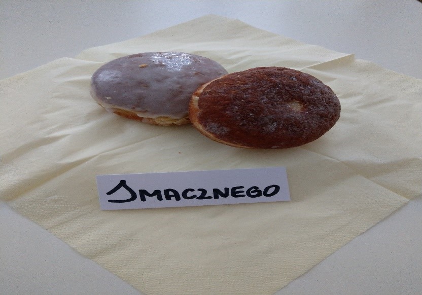 na fotografii widzimy dwa pączki ułożone na kremowej serwetce jeden pokryty lukrem a drugi czysty. Widoczny jest również na białej kartce napis „smacznego”