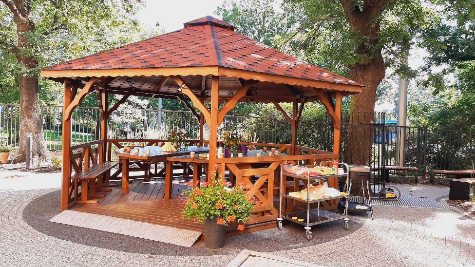 Na zdjęciu drewniana altana Dziennego Domu Pomocy. Jest to miejsce wspólnego grillowania. Po prawej stronie altany wózek gastronomiczny z przygotowanymi sałatkami , napojami 