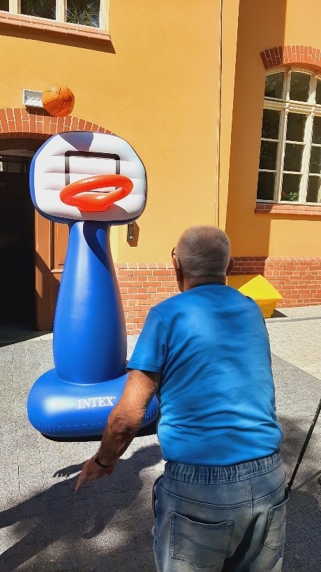 Na zdjęciu kosz do gry w koszykówkę niebiesko-białym i czerwonym kolorze oraz rzucający piłkę pan Jan. Na drugim planie Dzienny dom Pomocy przy Skoczylasa 8