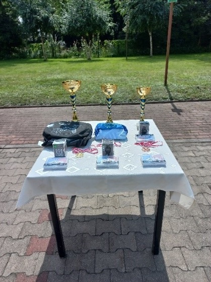Zdjęcie nr.7 Zdjęcie przedstawia puchary oraz nagrody za 1,2 i 3 miejsce. Leżą one na stole, który jest pokryty obrus.