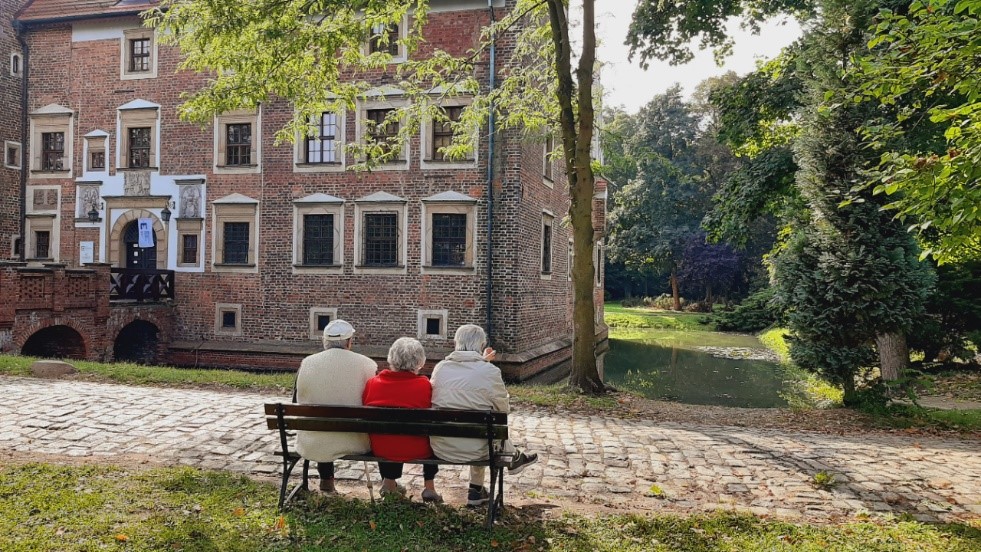 Zamek w Wojnowicach. Na pierwszym planie ławka na której odpoczywają nasi seniorzy. Na drugim planie zamek Wojnowicki, który zachwyca nas wyjątkowymi walorami architektonicznymi.