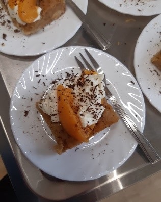 Szarlotka z bita śmietaną i brzoskwinią posypana czekoladą, tłem ciastka jest biały talerzyk. Szarlotka wykonana na zajęciach z kulinoterapii