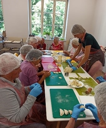 Zajęcia z kulinoterapii. na zdjęciu seniorzy lepiący pierogi ruskie. Biały stół, białe miseczki z farszem, kolorowe deski kuchenne na których układane są ulepione pierogi
