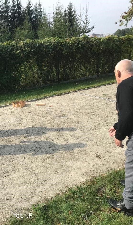 Na zdjęciu szare boisko z ustawionymi drewnianymi kręglami z prawej strony stojący mężczyzna w szaro czarnym dresie. W oddali drzewa i błękitne niebo
